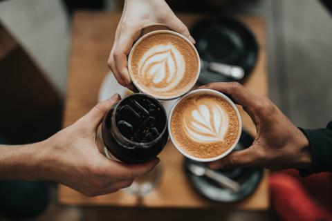 This image shows three hands holding drinks together in a toast. Two hands hold cups of coffee with beautiful latte art on top, and the third hand holds a glass of iced coffee. The background is softly blurred, suggesting a cozy café setting and a warm, social moment shared among friends.