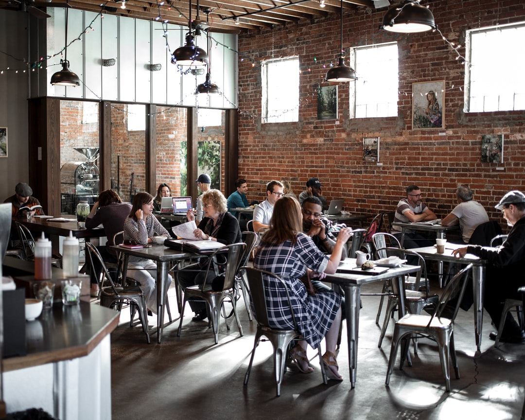 This image shows a lively, cozy cafe with a warm and rustic industrial design. The café has exposed brick walls, hanging pendant lights, and wooden ceiling beams. Customers are seated at metal tables, chatting, studying, or working on laptops, creating a casual, social atmosphere. Art and string lights add a creative touch to the ambiance. It feels like a welcoming spot for both work and conversation.