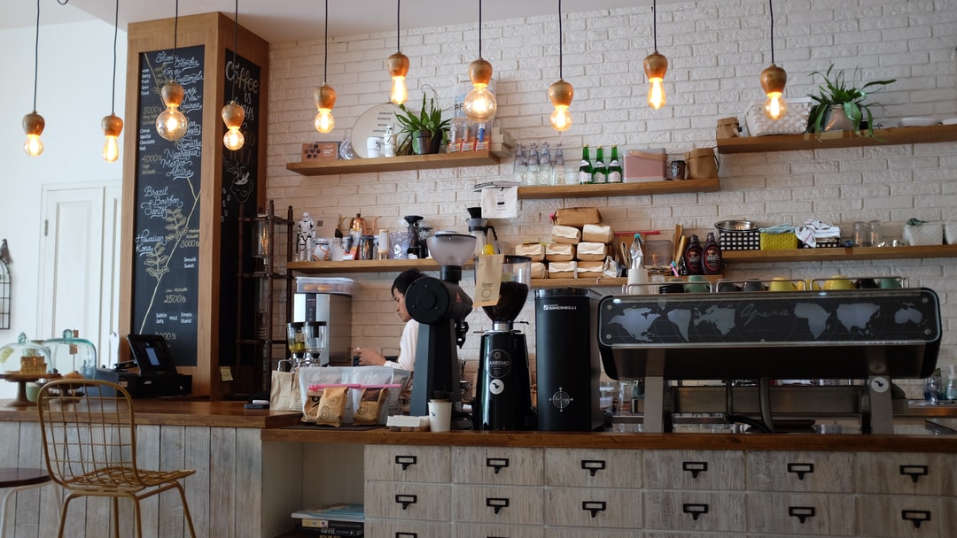 This image shows a modern, cozy coffee shop interior with a warm, inviting atmosphere. The counter is made of wood with a rustic finish, and above it hang several vintage-style light bulbs with exposed filaments, creating a soft glow. Behind the counter are shelves stocked with coffee bags, bottles, plants, and various equipment. A barista is preparing drinks, and the café features a clean white brick wall, a large espresso machine, and a chalkboard menu listing coffee options. The overall vibe is stylish and welcoming, perfect for coffee lovers.
